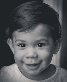 A close-up portrait of a young child with smooth skin, bright eyes, and a joyful expression. The child has short, neatly combed hair and is wearing small earrings. The image is in black and white, highlighting the textures and details of the child's face.