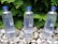 Three bottles of CBD Living Water are positioned among smooth pebbles with green foliage in the background. The bottles have blue caps and feature blue text with branding details. The setting is outdoors, providing a natural and refreshing backdrop.