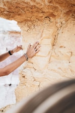 A person's hands are examining a textured, beige rock face, possibly for its geological features. The rock has visible layers and cracks, suggesting an ancient formation. Accessories like rings and bracelets are on the person's hand. The background shows more of the same rock formation.