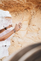 A person's hands are examining a textured, beige rock face, possibly for its geological features. The rock has visible layers and cracks, suggesting an ancient formation. Accessories like rings and bracelets are on the person's hand. The background shows more of the same rock formation.