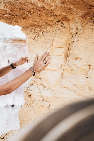 A person's hands are examining a textured, beige rock face, possibly for its geological features. The rock has visible layers and cracks, suggesting an ancient formation. Accessories like rings and bracelets are on the person's hand. The background shows more of the same rock formation.