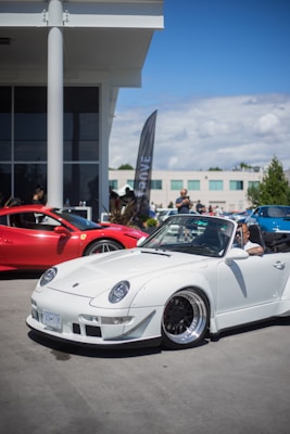A white convertible sports car is parked outdoors near a modern building. A red Ferrari is partially visible in the background. Several people are gathered around, and a tall flag with text is placed next to the cars.