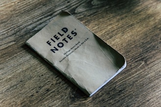 A small, worn notebook titled 'FIELD NOTES' lies on a wooden surface. The notebook cover is slightly creased, and the text specifies it as a 48-page memo book made in the USA using durable materials.