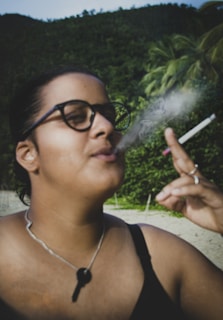 A person is outdoors, wearing glasses and smoking a cigarette, with smoke visibly emanating from it. The background features lush green foliage and palm trees, indicating a tropical setting.