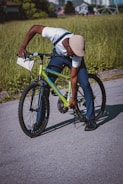 A person is crouching down next to a bright green bicycle, adjusting or inspecting the bike's stand. The background features a grassy area and a blurred view of buildings in the distance, suggesting an urban environment. The person is wearing a white shirt, dark pants, black shoes, and a light-colored hat.
