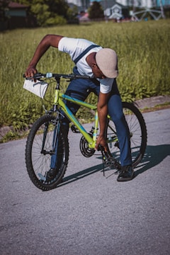 A person is crouching down next to a bright green bicycle, adjusting or inspecting the bike's stand. The background features a grassy area and a blurred view of buildings in the distance, suggesting an urban environment. The person is wearing a white shirt, dark pants, black shoes, and a light-colored hat.