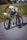 A person is crouching down next to a bright green bicycle, adjusting or inspecting the bike's stand. The background features a grassy area and a blurred view of buildings in the distance, suggesting an urban environment. The person is wearing a white shirt, dark pants, black shoes, and a light-colored hat.