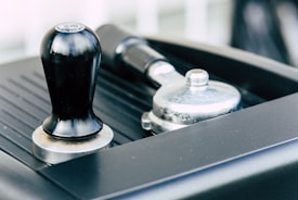 A close-up view of a black coffee tamper and a metal portafilter resting on a surface, possibly part of an espresso machine.