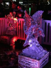 The scene features intricately carved ice sculptures illuminated by colored lights. The foreground displays a sculpture of a bird with outstretched wings, glowing with red and blue hues. In the background, another sculpture receives a red light projection, creating a vibrant atmosphere. The sculptures are placed on wooden platforms, with spectators visible in the darkened periphery.