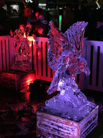 The scene features intricately carved ice sculptures illuminated by colored lights. The foreground displays a sculpture of a bird with outstretched wings, glowing with red and blue hues. In the background, another sculpture receives a red light projection, creating a vibrant atmosphere. The sculptures are placed on wooden platforms, with spectators visible in the darkened periphery.