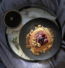 A breakfast setting featuring a stack of pancakes topped with berries and nuts on a dark plate. A cup of coffee is placed beside it on a marble tray. A small sprig of rosemary lies next to the plate. The tray is set on a grey, textured fabric.