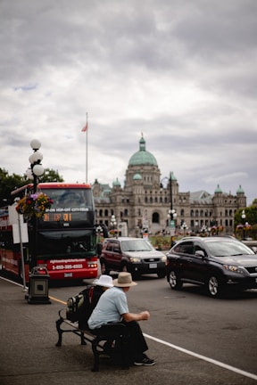 A city street scene with a red double-decker tour bus and several cars in front of a historic building with a green dome. Two individuals sit on a bench with backpacks, one wearing a blue shirt and the other a hat. Overcast skies with the Canadian flag visible on a pole.