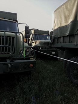 Military-style trucks parked closely together in a grassy area, featuring camouflage patterns and rugged exteriors. The setting appears to be outdoors under natural light.