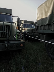 Military-style trucks parked closely together in a grassy area, featuring camouflage patterns and rugged exteriors. The setting appears to be outdoors under natural light.