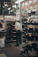 An elderly man wearing a hat and a shoulder bag is browsing shoes in a store aisle. Discount signs showing percentages off are hanging above him. Various shoes are displayed on metal racks and shelves, and brands such as Adidas and Reebok are visible on store banners.