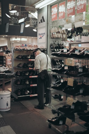 An elderly man wearing a hat and a shoulder bag is browsing shoes in a store aisle. Discount signs showing percentages off are hanging above him. Various shoes are displayed on metal racks and shelves, and brands such as Adidas and Reebok are visible on store banners.
