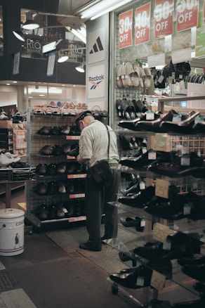 An elderly man wearing a hat and a shoulder bag is browsing shoes in a store aisle. Discount signs showing percentages off are hanging above him. Various shoes are displayed on metal racks and shelves, and brands such as Adidas and Reebok are visible on store banners.