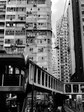 A densely packed urban environment featuring tall residential buildings with numerous windows and balconies. An elevated pedestrian walkway crosses the scene, with people visible beneath it. The architecture is typical of a busy city setting, characterized by straight lines and uniform construction. The atmosphere exudes a sense of crowdedness and urban life.