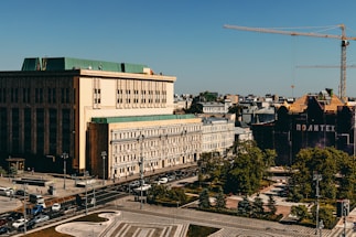 An urban landscape featuring a large building with a green roof and a smaller building beside it. A construction crane towers over another dark building with the word 'Политех.' The scene is busy with vehicles on the roads and trees lining the sidewalks, set against a clear blue sky.