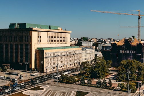 An urban landscape featuring a large building with a green roof and a smaller building beside it. A construction crane towers over another dark building with the word 'Политех.' The scene is busy with vehicles on the roads and trees lining the sidewalks, set against a clear blue sky.