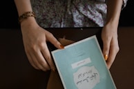 A pair of hands with painted fingernails hold a book with a blue cover. The text on the book appears to be in a foreign script. The person is wearing a floral-patterned top and a brown beaded bracelet. The book is placed on a brown surface, and a wallet or folder is partially visible beneath it.