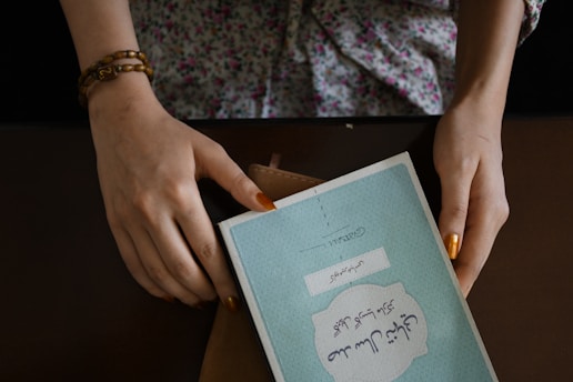 A pair of hands with painted fingernails hold a book with a blue cover. The text on the book appears to be in a foreign script. The person is wearing a floral-patterned top and a brown beaded bracelet. The book is placed on a brown surface, and a wallet or folder is partially visible beneath it.