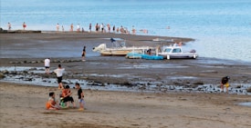 A beach scene features groups of people enjoying various activities. Several children are gathered in a circle on the sand, while others walk along the shore. Two boats are docked in the shallow water, surrounded by adults and children wading in the sea. The overall setting is relaxed and casual, with a bright blue sky in the background.