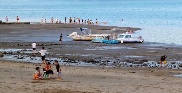 A beach scene features groups of people enjoying various activities. Several children are gathered in a circle on the sand, while others walk along the shore. Two boats are docked in the shallow water, surrounded by adults and children wading in the sea. The overall setting is relaxed and casual, with a bright blue sky in the background.