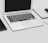 A MacBook Air laptop is open on a desk. Adjacent to the laptop are a closed book and a smartphone, both placed on the left side of the image. To the right of the laptop is a wireless mouse partially visible. The surface is sleek and minimalistic, suggesting a clean and organized workspace.
