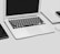 A MacBook Air laptop is open on a desk. Adjacent to the laptop are a closed book and a smartphone, both placed on the left side of the image. To the right of the laptop is a wireless mouse partially visible. The surface is sleek and minimalistic, suggesting a clean and organized workspace.