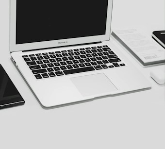 A MacBook Air laptop is open on a desk. Adjacent to the laptop are a closed book and a smartphone, both placed on the left side of the image. To the right of the laptop is a wireless mouse partially visible. The surface is sleek and minimalistic, suggesting a clean and organized workspace.