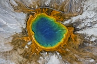 An aerial view of a vibrant geothermal spring displaying concentric rings of colors, with a deep blue center surrounded by a gradient of green, yellow, and orange hues. The area around the spring features contrasting grey and brown earth tones, and there is a walkway or boardwalk visible near the edge.