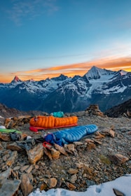 A scenic mountain landscape with snow-capped peaks in the background under a vibrant sunset sky. In the foreground, there are two sleeping bags, one orange and one blue, placed on rocky ground. Camping equipment and gear are scattered around the area, suggesting an outdoor camping setup.