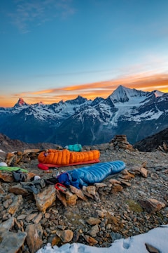 A scenic mountain landscape with snow-capped peaks in the background under a vibrant sunset sky. In the foreground, there are two sleeping bags, one orange and one blue, placed on rocky ground. Camping equipment and gear are scattered around the area, suggesting an outdoor camping setup.