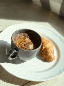 A warm cup of coffee next to a flaky croissant on a white ceramic plate near a sunlit window.