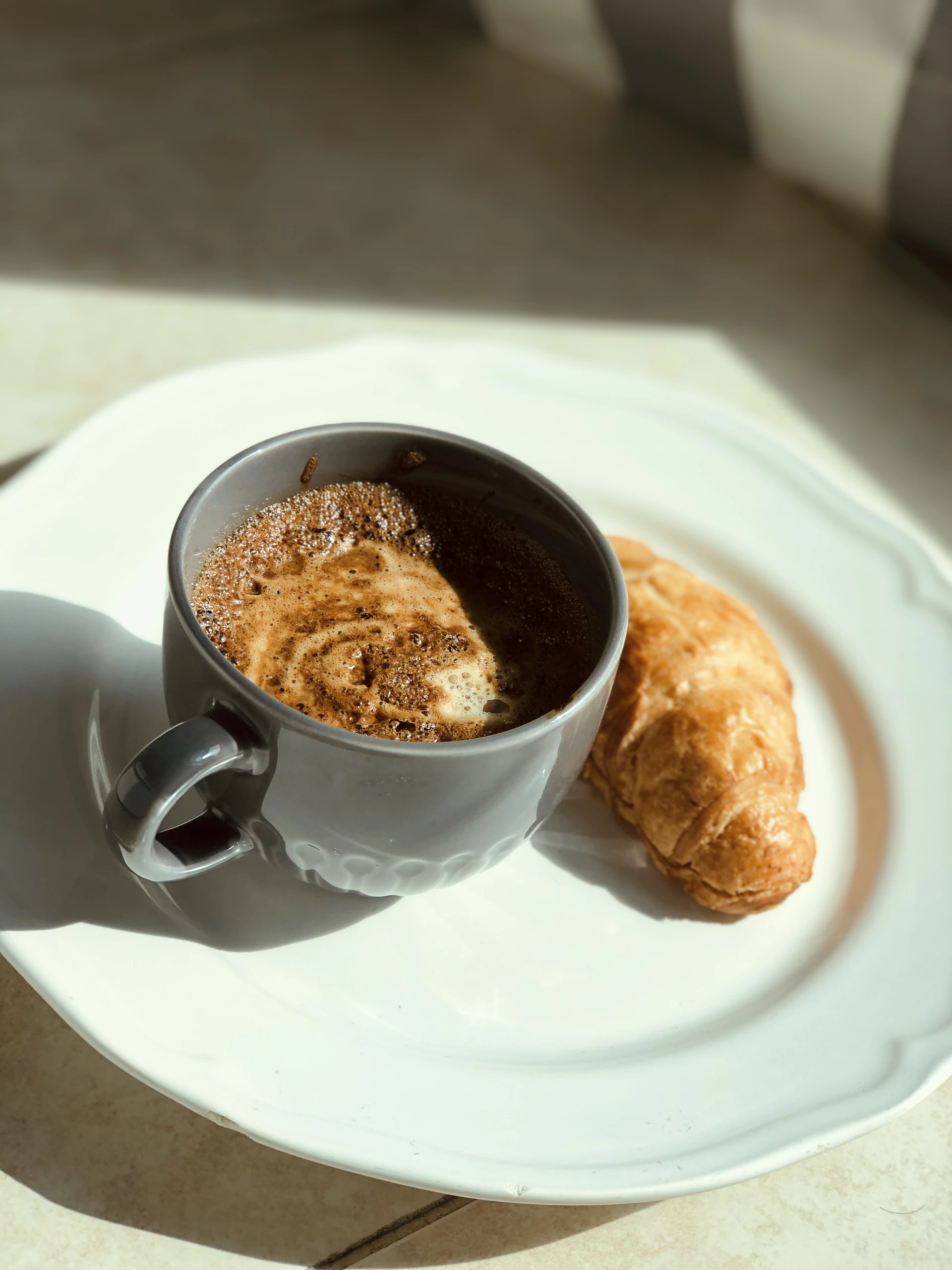 A close-up shot of a glossy, gold-accented coffee cup steaming beside a freshly baked croissant on a sleek black marble table, bathed in warm morning light.