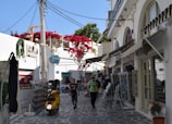 A picturesque alley in a Mediterranean town, featuring whitewashed buildings with arched windows and doors. Bright red flowers cascade over a balcony. A yellow scooter is parked on the cobblestone path, and two people are walking along the street, surrounded by various shops displaying postcards and other items.