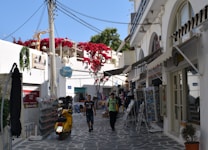 A picturesque alley in a Mediterranean town, featuring whitewashed buildings with arched windows and doors. Bright red flowers cascade over a balcony. A yellow scooter is parked on the cobblestone path, and two people are walking along the street, surrounded by various shops displaying postcards and other items.