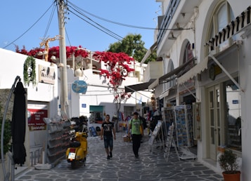 A picturesque alley in a Mediterranean town, featuring whitewashed buildings with arched windows and doors. Bright red flowers cascade over a balcony. A yellow scooter is parked on the cobblestone path, and two people are walking along the street, surrounded by various shops displaying postcards and other items.