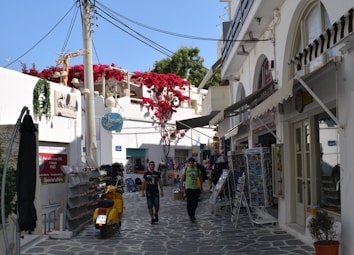 A picturesque alley in a Mediterranean town, featuring whitewashed buildings with arched windows and doors. Bright red flowers cascade over a balcony. A yellow scooter is parked on the cobblestone path, and two people are walking along the street, surrounded by various shops displaying postcards and other items.