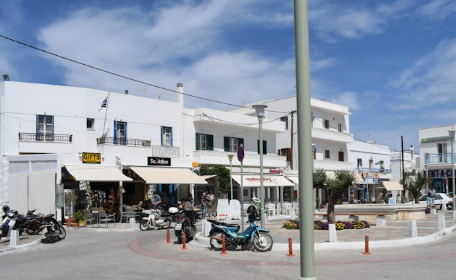 A sunlit street scene in a small town featuring white-washed buildings with balconies. Several cafes and shops are present with outdoor seating and people enjoying the day. Motorcycles and scooters are parked nearby, and there are a few trees and planters with flowers around the square.