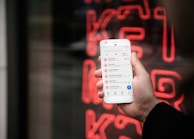 A hand holding a smartphone displaying a list of recent calls, mostly marked as spam or telemarketing. In the background, there is a neon red sign with indistinct letters or symbols.