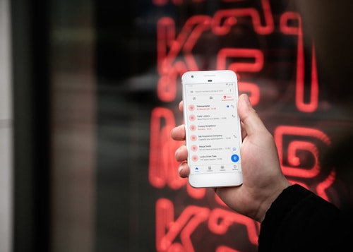 A hand holding a smartphone displaying a list of recent calls, mostly marked as spam or telemarketing. In the background, there is a neon red sign with indistinct letters or symbols.