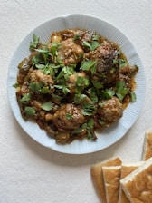 A white plate filled with cooked meatballs garnished with fresh green herbs, surrounded by a tomato-based sauce, possibly including vegetables. The plate is placed on a textured white surface, and slices of flatbread are positioned nearby.