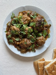 A white plate filled with cooked meatballs garnished with fresh green herbs, surrounded by a tomato-based sauce, possibly including vegetables. The plate is placed on a textured white surface, and slices of flatbread are positioned nearby.