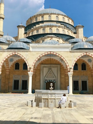A large mosque featuring intricate Islamic architecture with multiple domes, arches, and patterned tiles. A central water fountain, used for ablutions, is in the courtyard, with a person seated nearby. The mosque's façade displays a blend of stone and blue tiles, and the sky above is clear and bright.