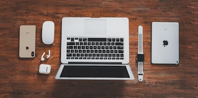 A collection of Apple devices arranged on a wooden surface, including a laptop, smartphone, tablet, smartwatch, wireless earbuds, and a mouse. The devices are neatly aligned, showcasing modern digital technology.