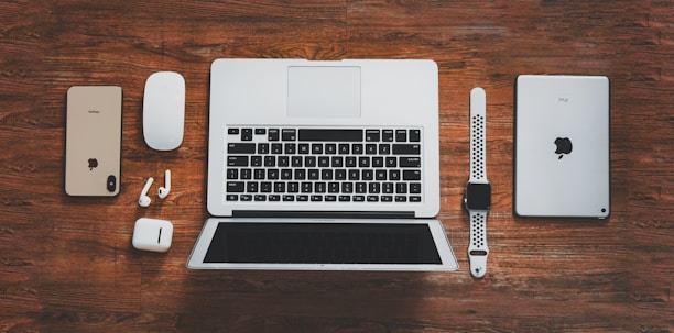 A collection of Apple devices arranged on a wooden surface, including a laptop, smartphone, tablet, smartwatch, wireless earbuds, and a mouse. The devices are neatly aligned, showcasing modern digital technology.