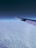 An airplane wing extends over a blanket of fluffy white clouds, with a gradient sky transitioning from dark blue at the top to lighter hues near the horizon.