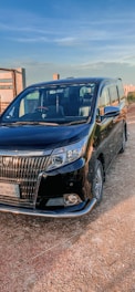 A black minivan parked on a dirt road, with a clear blue sky in the background. The vehicle is angled slightly to the right, revealing its sleek design and shiny exterior. The front grille is prominent, and the license plate is visible. The surroundings include a fence and open land, suggesting a rural or suburban setting.